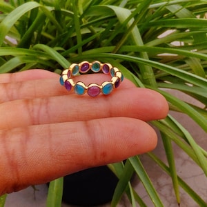 May include: A gold band ring featuring a continuous row of alternating round, cabochon-cut gemstones in vibrant shades of turquoise blue, deep magenta pink, and purple. The polished gold metal setting holds each individual stone securely in place.