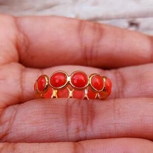 May include: A gold band ring featuring a continuous row of small, round, vibrant orange-red cabochon stones set in individual metal bezels that encircle the entire circumference of the jewelry piece, creating a uniform and repeating circular pattern.