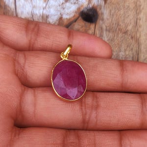 May include: A gold pendant with a large, faceted, oval-shaped ruby gemstone. The ruby is a deep red color and is set in a gold bezel. The pendant is held in a hand against a wooden background.