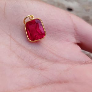 Puede incluir: Un colgante dorado con una gema rectangular de color rojo intenso. El colgante tiene un pequeño lazo en la parte superior para una cadena. La gema está engastada en un marco dorado.