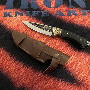 May include: A decorative knife with a detailed blade and a black handle, resting beside a brown leather sheath. The blade features an ornate design. The text "IRON KNIFE ART" is visible in the background.