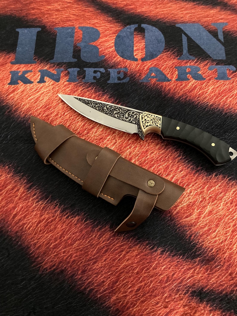 May include: A decorative knife with a detailed blade and a black handle, resting beside a brown leather sheath. The blade features an ornate design. The text "IRON KNIFE ART" is visible in the background.