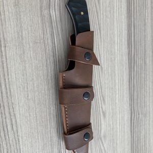 May include: A black-handled knife with a silver blade is sheathed in a brown leather case. The case has three straps with black snap closures. The knife and sheath are set against a light wood grain background.