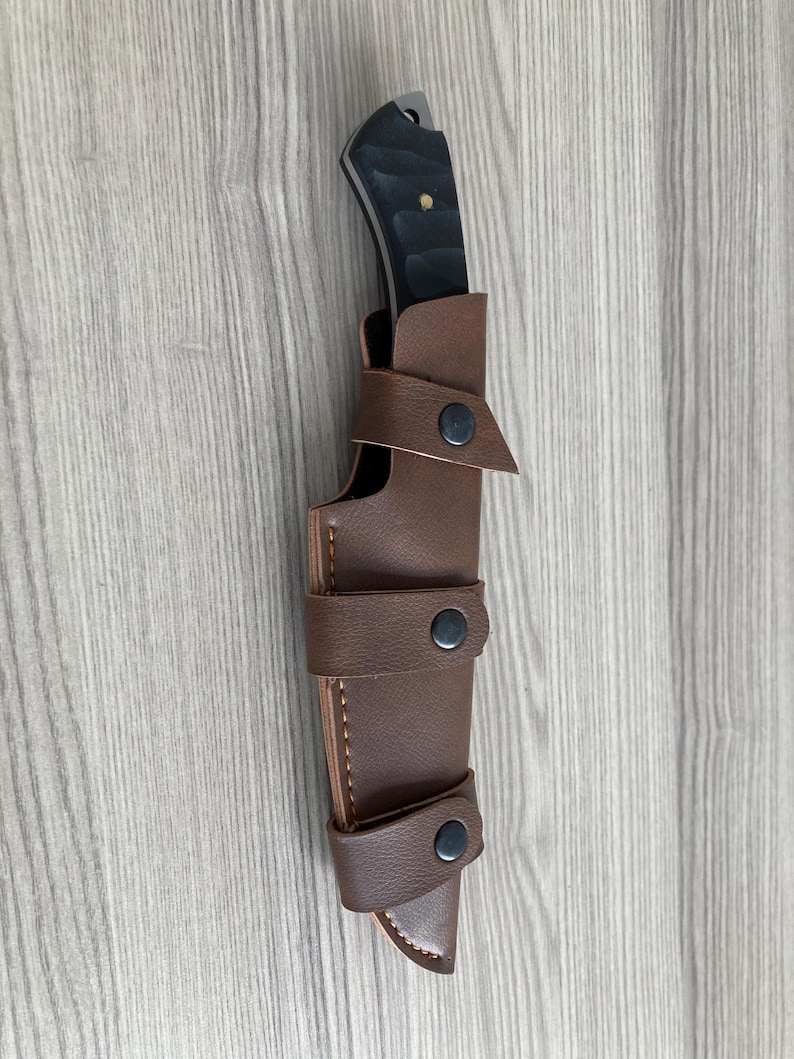 May include: A black-handled knife with a silver blade is sheathed in a brown leather case. The case has three straps with black snap closures. The knife and sheath are set against a light wood grain background.