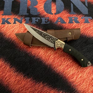 May include: An ornate knife with a silver blade and a gold-colored handle, resting on a brown leather sheath. The handle is black with gold accents. The background features the text "IRON KNIFE ART" over a tiger stripe pattern.