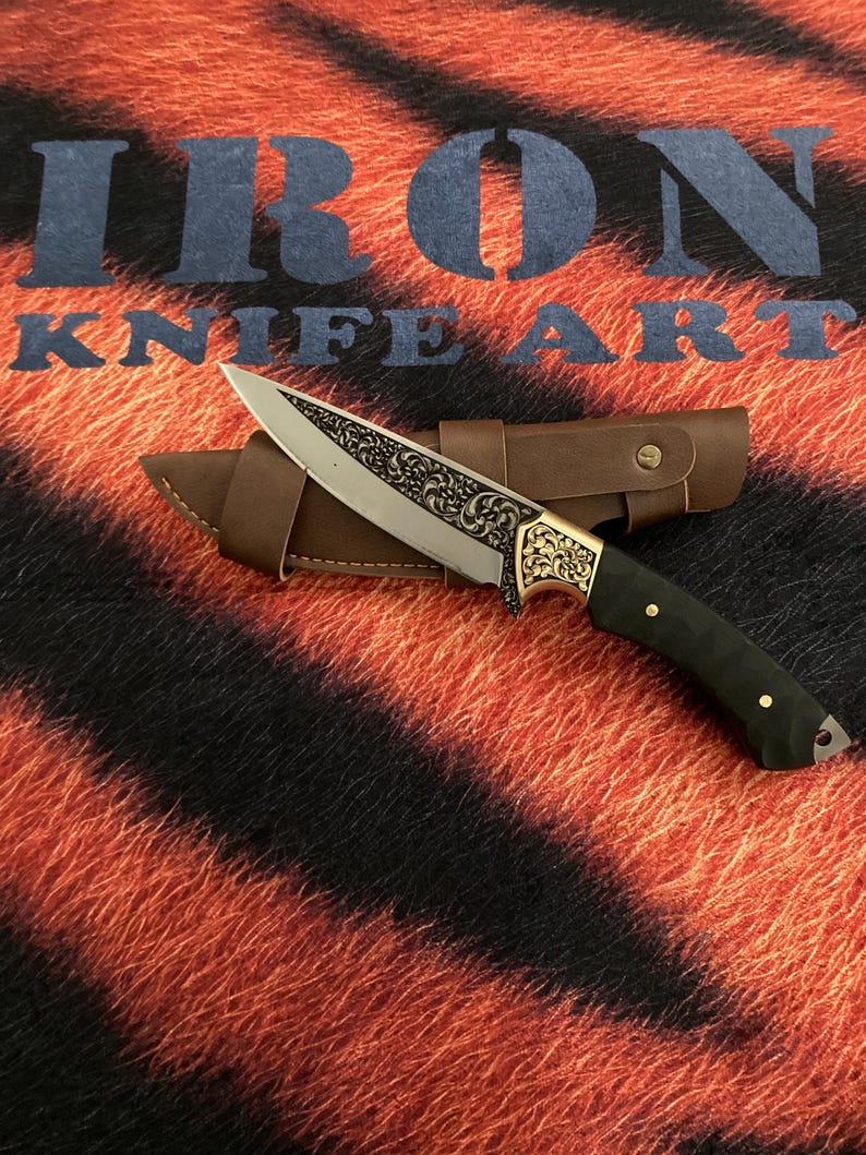 May include: An ornate knife with a silver blade and a gold-colored handle, resting on a brown leather sheath. The handle is black with gold accents. The background features the text "IRON KNIFE ART" over a tiger stripe pattern.
