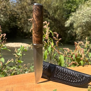 Könnte beinhalten: Ein feststehendes Messer mit einer polierten Metallklinge und einem Griff aus dunkel gemasertem Holz, ergänzt durch eine silberfarbene Zwinge. Neben dem Messer liegt eine schwarze Lederscheide mit einem geprägten, geflochtenen Muster.