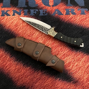 May include: A decorative knife with a silver blade and ornate gold detailing on the handle, accompanied by a brown leather sheath. The knife features a black handle and is displayed on a textured background with the text "IRON KNIFE ART".