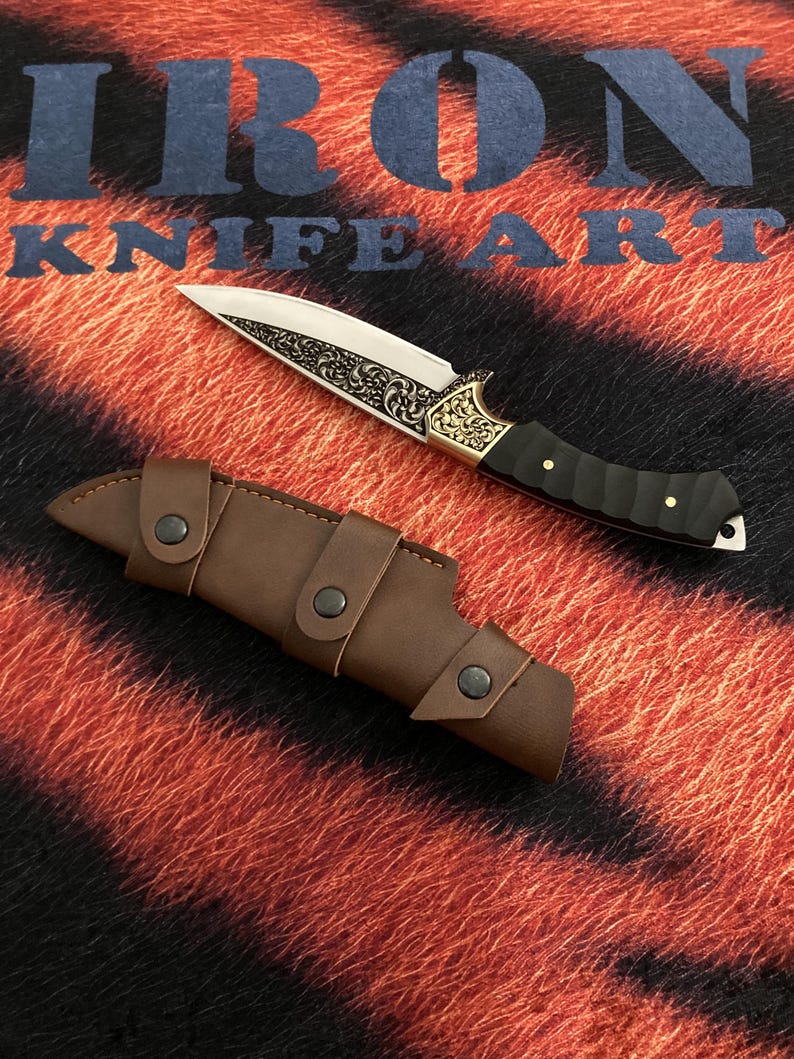 May include: A decorative knife with a silver blade and ornate gold detailing on the handle, accompanied by a brown leather sheath. The knife features a black handle and is displayed on a textured background with the text "IRON KNIFE ART".
