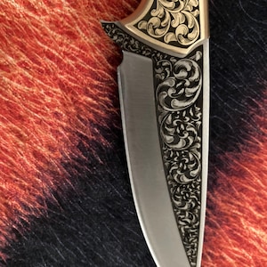May include: Close-up of a decorative knife with a silver blade and ornate gold-colored handle. The handle features intricate floral engravings. The knife's design is symmetrical, with detailed patterns along the blade's edge and handle.