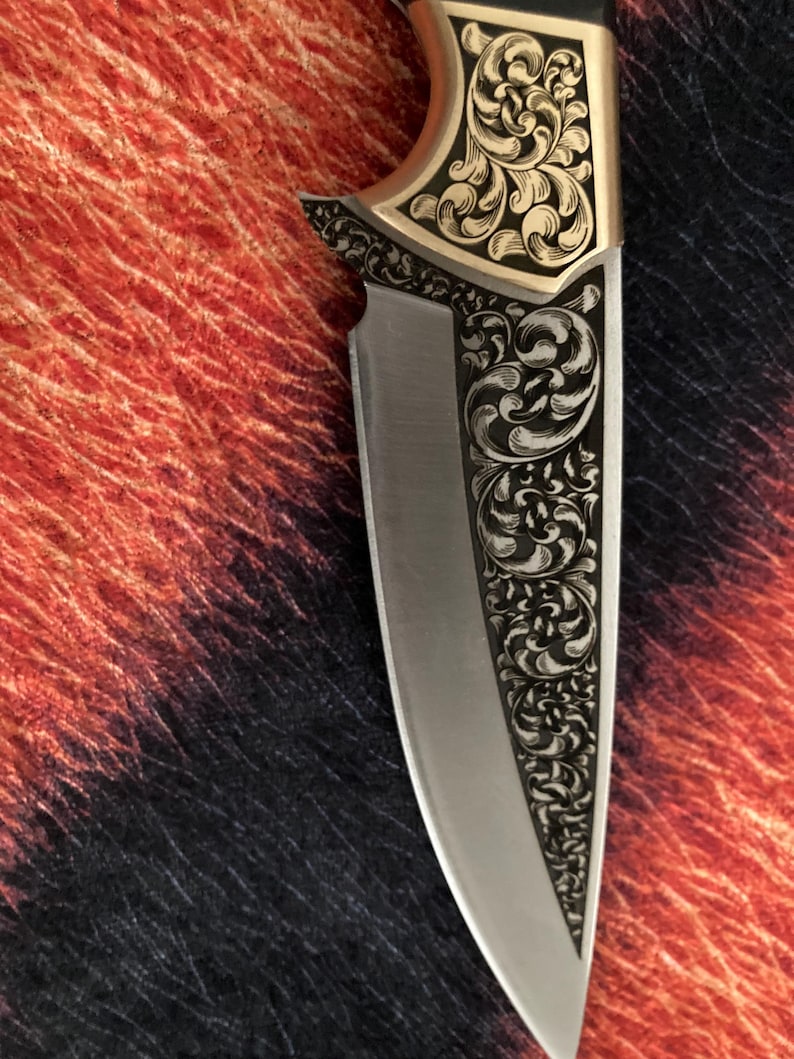 May include: Close-up of a decorative knife with a silver blade and ornate gold-colored handle. The handle features intricate floral engravings. The knife's design is symmetrical, with detailed patterns along the blade's edge and handle.