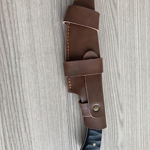 May include: A brown leather knife sheath with a knife inside. The sheath has a strap and button closure. The knife handle is black with a textured design. The knife blade is silver.