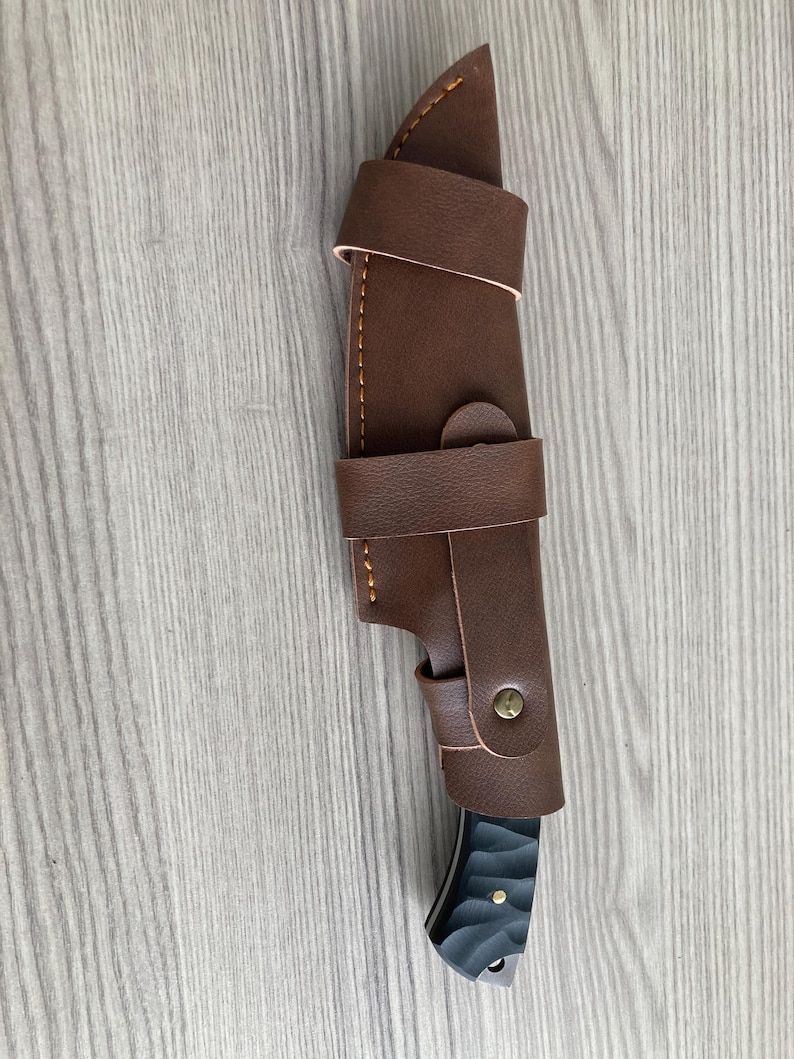 May include: A brown leather knife sheath with a knife inside. The sheath has a strap and button closure. The knife handle is black with a textured design. The knife blade is silver.