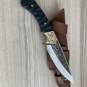 May include: A handcrafted hunting knife with a black textured handle and a decorative gold-colored bolster. The blade is silver with an engraved floral pattern. It is housed in a brown leather sheath.