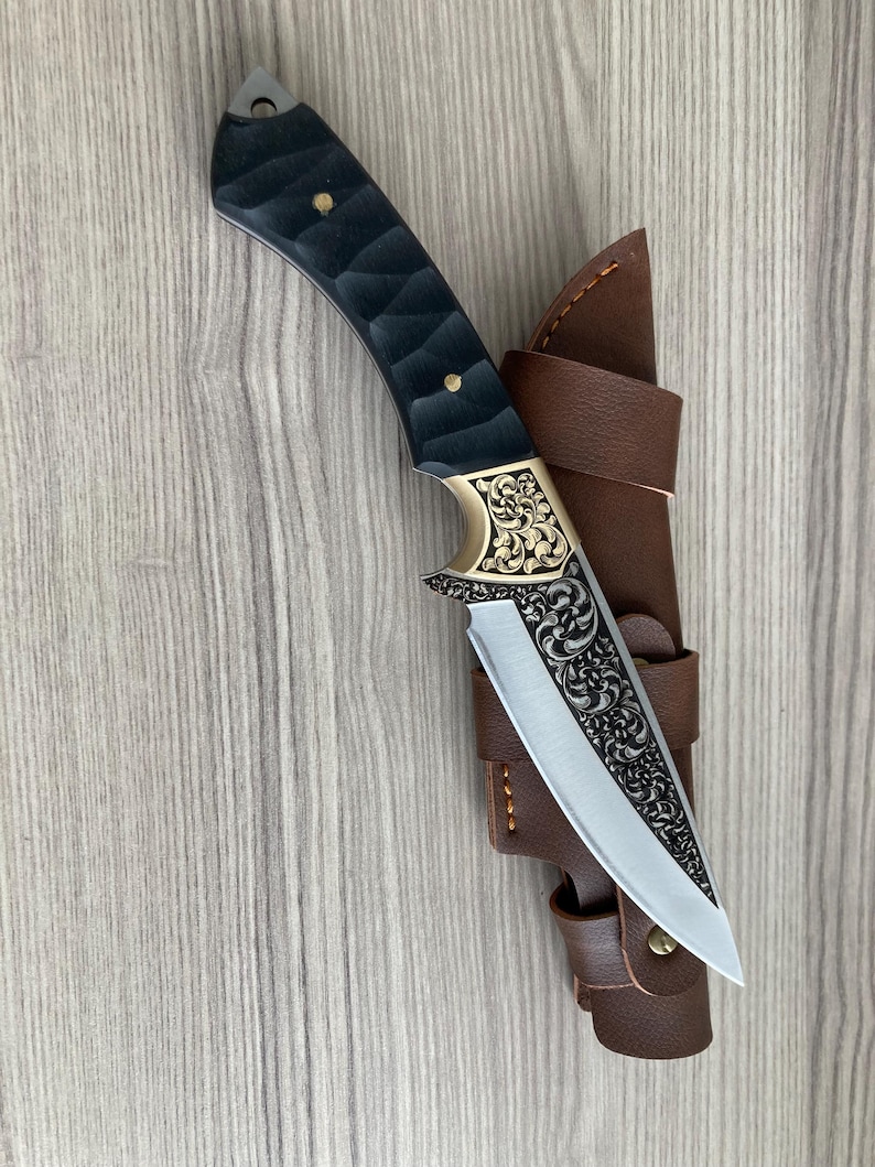 May include: A handcrafted hunting knife with a black textured handle and a decorative gold-colored bolster. The blade is silver with an engraved floral pattern. It is housed in a brown leather sheath.