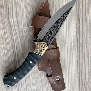 May include: A decorative hunting knife with a silver blade featuring an ornate floral design. The handle is black with gold accents and brass rivets. It comes with a brown leather sheath.