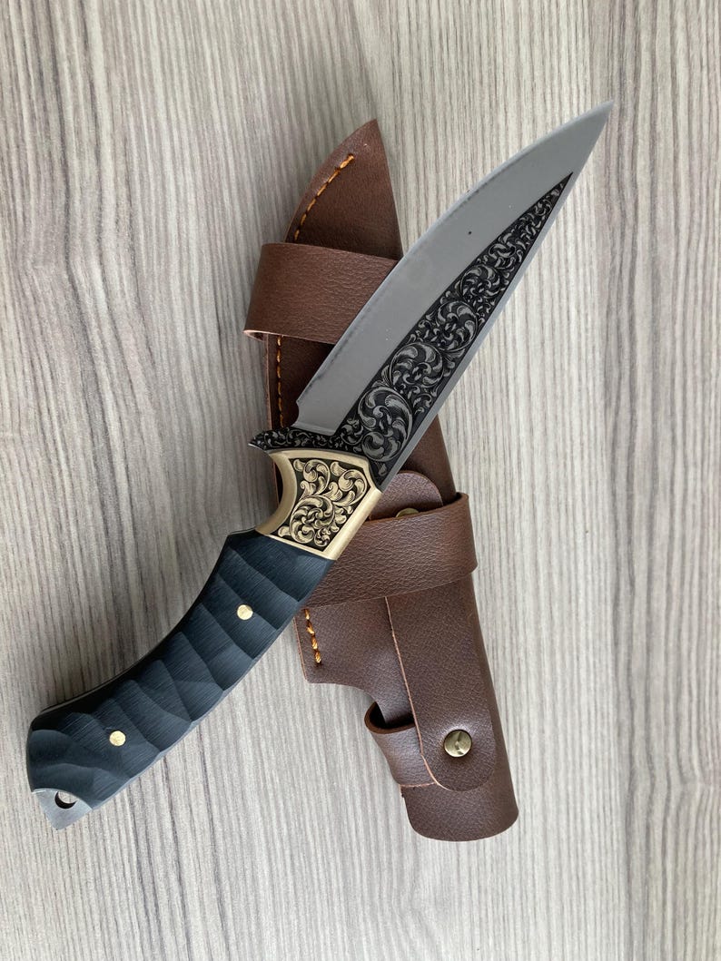 May include: A decorative hunting knife with a silver blade featuring an ornate floral design. The handle is black with gold accents and brass rivets. It comes with a brown leather sheath.