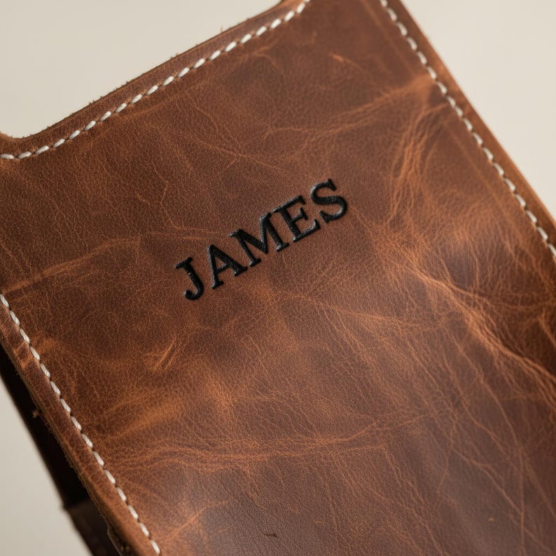 以下が含まれることがあります： 黒字で「JAMES」と刻印された茶色の革製携帯電話ケースのクローズアップ。ケースの縁には白いステッチが施され、革の自然な風合いを活かした、使い古したような仕上がりになっています。