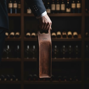 以下が含まれることがあります： 茶色の革製ワインボトルキャリー、持ち手付き。人が持っています。前面には「JAMES」の文字がエンボス加工されています。背景にはワインセラーとボトルが写っています。