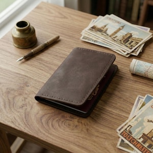 May include: A brown leather passport holder sits open on a wooden surface, next to a vintage inkwell, pen, and postcards. The passport holder has a stitched border and a textured surface. A stack of postcards with European landmarks is visible.