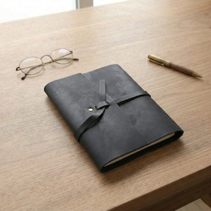 Puede incluir: Un diario de cuero negro con cierre de lazo y un botón de latón. Unas gafas redondas y un bolígrafo dorado y de madera descansan sobre una superficie de madera junto al diario.