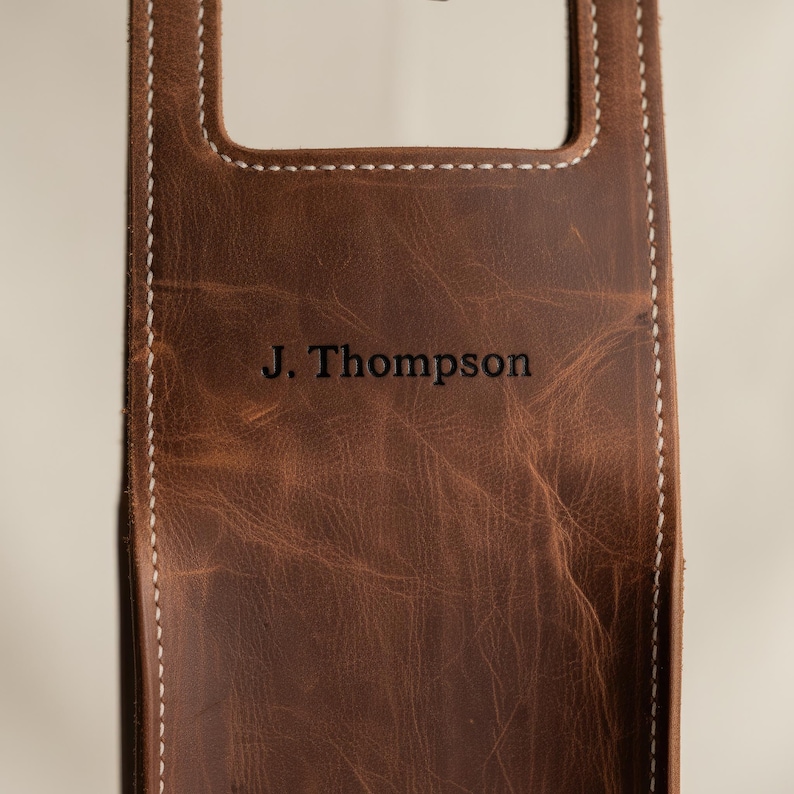 以下が含まれることがあります： 「J. Thompson」の刻印が入った茶色の革製品のクローズアップ。革は使い古したような外観で、縁には白いステッチが施されています。持ち手が付いた長方形の形をしています。
