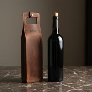 以下が含まれることがあります： 茶色の革製ワインボトルキャリー。ハンドルと前面に「JAMES」のエンボス加工が施されています。隣にはコルク栓付きの濃い色のガラス製ワインボトルがあります。キャリーとボトルは、大理石の表面に置かれています。