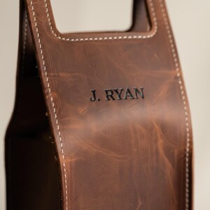以下が含まれることがあります： 茶色の革製ワインボトルキャリー。白い糸でステッチされ、「J. RYAN」のエンボス加工が施されています。革は使い古したような外観です。