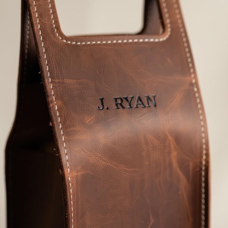 以下が含まれることがあります： 茶色の革製ワインボトルキャリー。白い糸でステッチされ、「J. RYAN」のエンボス加工が施されています。革は使い古したような外観です。