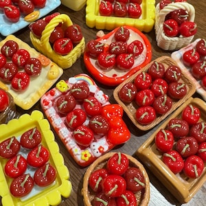 Könnte beinhalten: Eine Sammlung von Miniatur-Kirsch-Anhängern in Rot mit grünen Stielen, präsentiert in verschiedenen kleinen Behältern. Dazu gehören Körbe, Tabletts und Schalen in Farben wie Gelb, Blau und Weiß. Die Kirschen haben eine glänzende Oberfläche.