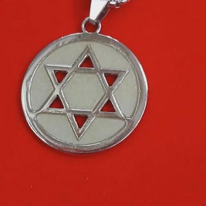 Può includere: Una collana con pendente color argento con un design a Stella di David. Il pendente ha una cornice circolare con una stella a sei punte ritagliata al centro. La stella è riempita con un materiale di colore chiaro. Anche la catena della collana è argentata.