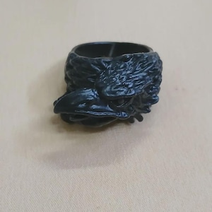 May include: A black, raven-shaped ring holder. The ring holder is made of a dark, glossy material and features a detailed carving of a raven's head, including its beak and feathers. The ring holder is designed to hold rings and other small jewelry items.
