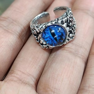 May include: A silver-colored ring featuring a large, round, blue eye design. The eye has intricate details and a dark pupil. The ring has a textured, organic design around the eye, and the band is adjustable.