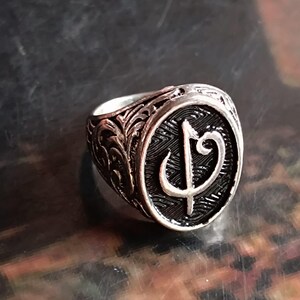 Puede incluir: Un anillo plateado con un dise&ntilde;o grabado. El anillo presenta una cara circular con un s&iacute;mbolo estilizado en el centro. La banda est&aacute; detallada con patrones ornamentados. El anillo est&aacute; sobre un fondo oscuro.