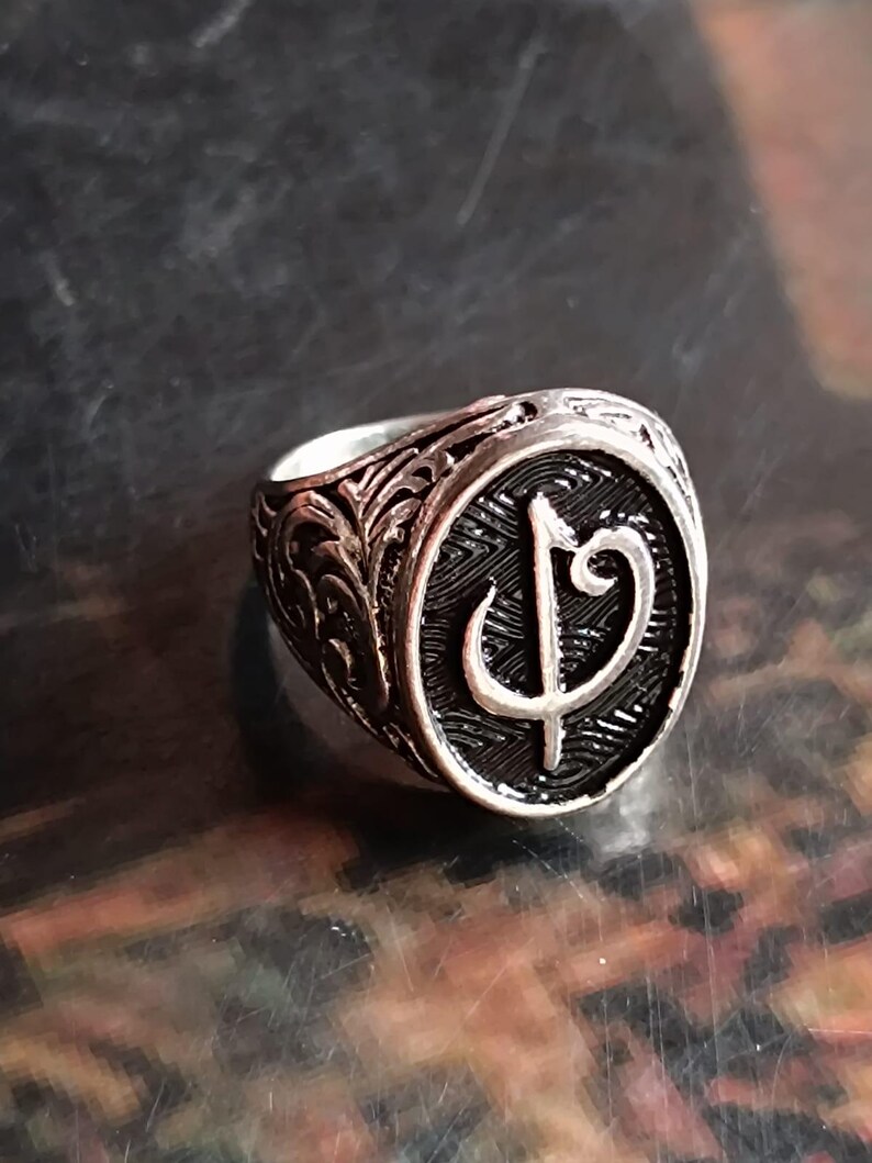 Puede incluir: Un anillo plateado con un dise&ntilde;o grabado. El anillo presenta una cara circular con un s&iacute;mbolo estilizado en el centro. La banda est&aacute; detallada con patrones ornamentados. El anillo est&aacute; sobre un fondo oscuro.