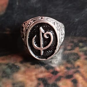 Puede incluir: Anillo plateado con un centro ovalado negro que presenta un s&iacute;mbolo estilizado. El anillo tiene un dise&ntilde;o ornamentado y grabado alrededor de la banda y el centro ovalado. El anillo es probablemente de metal.