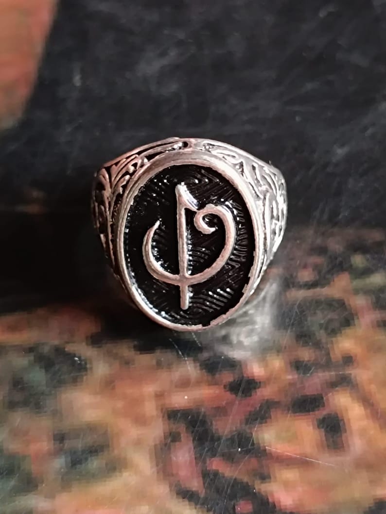 Puede incluir: Anillo plateado con un centro ovalado negro que presenta un s&iacute;mbolo estilizado. El anillo tiene un dise&ntilde;o ornamentado y grabado alrededor de la banda y el centro ovalado. El anillo es probablemente de metal.