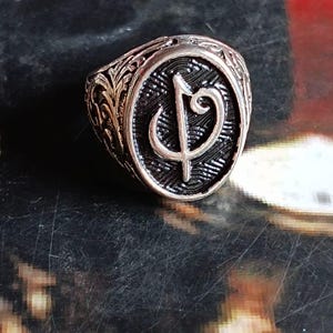 Puede incluir: Anillo plateado con una pieza central ovalada con un s&iacute;mbolo estilizado. El anillo tiene una banda ornamentada, grabada con un patr&oacute;n floral detallado. El s&iacute;mbolo se encuentra sobre un fondo oscuro y texturizado.