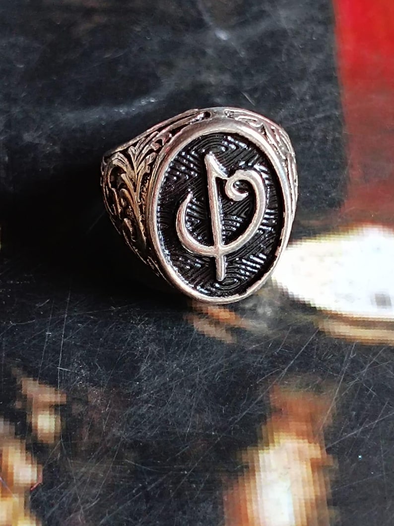 Puede incluir: Anillo plateado con una pieza central ovalada con un s&iacute;mbolo estilizado. El anillo tiene una banda ornamentada, grabada con un patr&oacute;n floral detallado. El s&iacute;mbolo se encuentra sobre un fondo oscuro y texturizado.