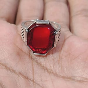 May include: A silver-toned ring featuring a large, rectangular, deep red gemstone. The ring has a detailed band with textured patterns on the sides. The gemstone is set in a silver bezel, creating a striking contrast.