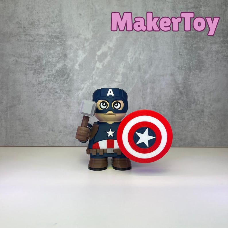 May include: A Captain America figurine with a shield and hammer. The figurine is wearing a blue helmet with a white "A" and a star on the chest. The shield is red, white, and blue. The text "MakerToy" is in pink.