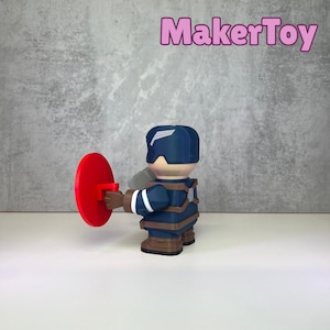 May include: A Captain America figurine with a red shield. The figurine is wearing a blue and brown uniform and helmet. The text "MakerToy" is in pink in the upper right corner.