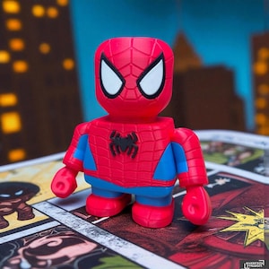 May include: A red and blue Spider-Man figurine with a black spider emblem on the chest. The figure has white eye details and is posed on a comic book page. The figure is a stylized, blocky design.