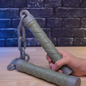 Op de afbeelding: Olijfgroene nunchaku's met een ketting en getextureerde handvatten. De handvatten worden door een hand vastgehouden. De tekst "KID FRIENDLY BUILDS!" staat bovenaan de afbeelding.