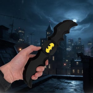 May include: A black Batarang, a Batman-themed weapon, featuring a yellow and black emblem. The image shows the Batarang against a backdrop of a city at night.