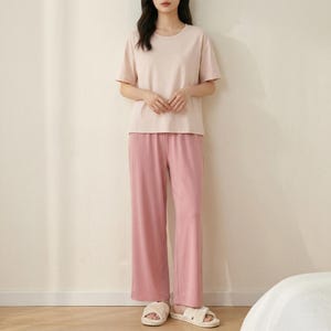 May include: A two-piece lounge set consisting of a short-sleeved, crew-neck t-shirt in a pale blush pink and matching wide-leg trousers in a slightly darker shade of pink, paired with light-colored, open-toed slide sandals.