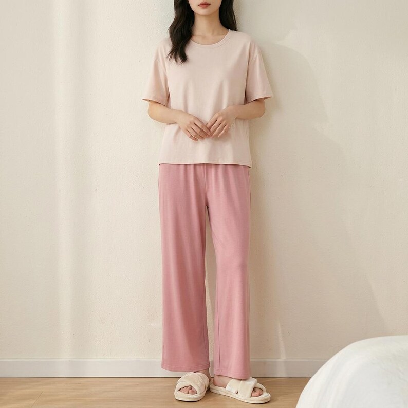 May include: A two-piece lounge set consisting of a short-sleeved, crew-neck t-shirt in a pale blush pink and matching wide-leg trousers in a slightly darker shade of pink, paired with light-colored, open-toed slide sandals.