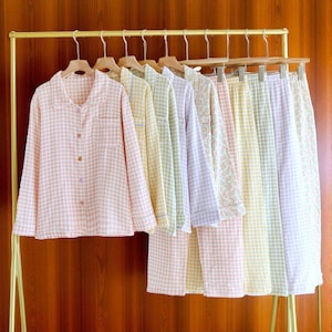 Könnte beinhalten: Eine Kollektion pastellfarbener Pyjama-Sets, die an einer goldenen Kleiderstange hängen. Die Sets umfassen langärmlige Hemden mit Knöpfen und passende Hosen in rosa, gelben, grünen und lavendelfarbenen Vichy-Mustern sowie einen Blumendruck.