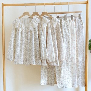 Peut inclure: Plusieurs ensembles de pyjamas blancs à motifs floraux sont suspendus à un portant en bois. Les hauts ont un col rond à volants et des manches longues. Les pantalons sont longs et amples, avec un motif floral assorti. Les pyjamas sont en tissu léger.