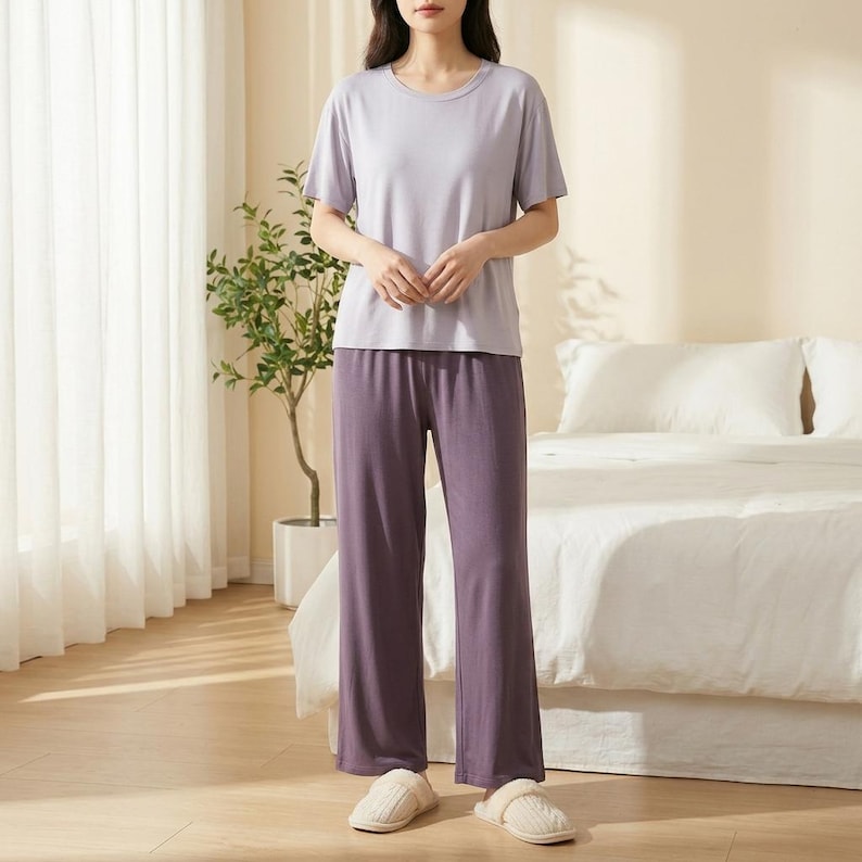 May include: A two-piece lounge set consisting of a short-sleeved, light lavender crew-neck t-shirt and matching dark purple wide-leg trousers. The set is paired with beige, closed-toe, textured slip-on slippers with a rounded front design.